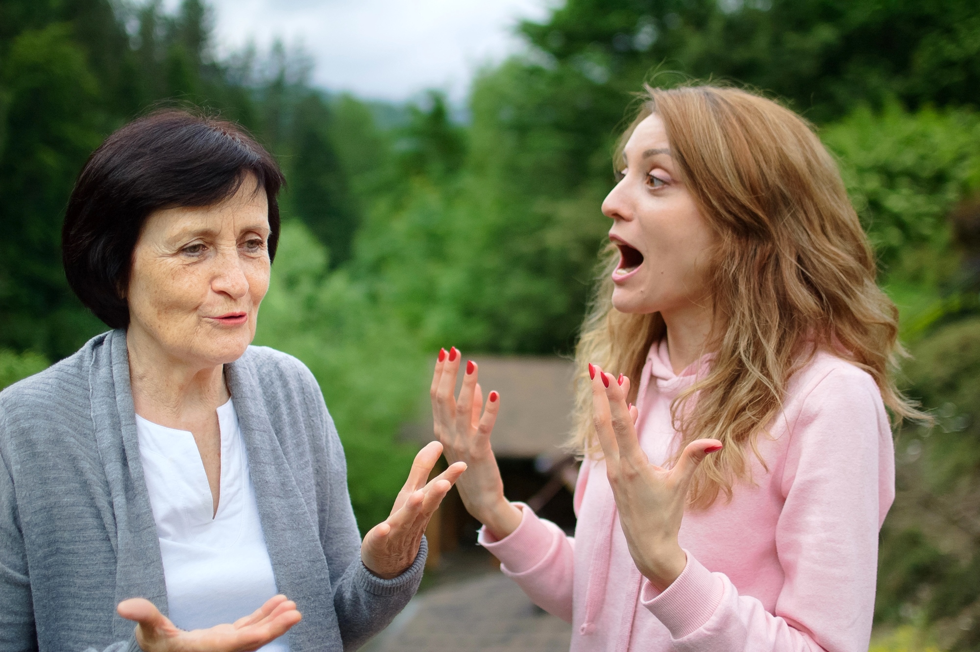 Two women having an animated conversation in an outdoor setting. The older woman, wearing a gray cardigan, gestures with her hands, while the younger woman in a pink hoodie appears surprised, with hands raised. Lush greenery is in the background.