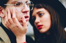 A young woman with red lipstick gently holds the face of a young man wearing glasses. The background is blurred, highlighting their expressions. The atmosphere is intimate and thoughtful.