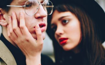 A young woman with red lipstick gently holds the face of a young man wearing glasses. The background is blurred, highlighting their expressions. The atmosphere is intimate and thoughtful.