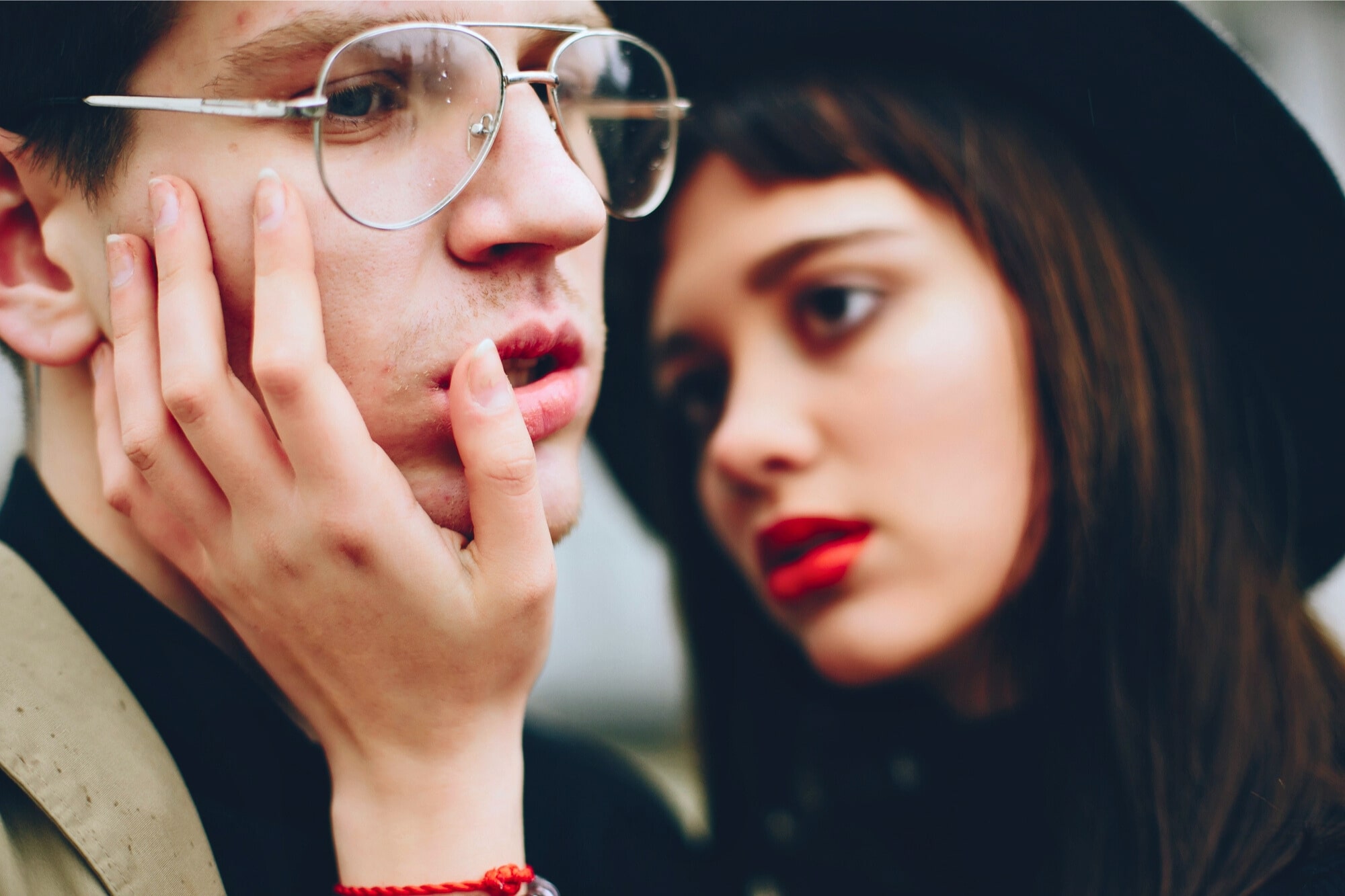 A young woman with red lipstick gently holds the face of a young man wearing glasses. The background is blurred, highlighting their expressions. The atmosphere is intimate and thoughtful.