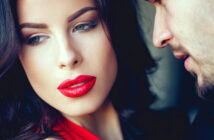 A woman with dark hair and red lipstick looks away thoughtfully while a man leans in close to whisper something, partially visible. The scene conveys an intimate and possibly intense moment between the two.