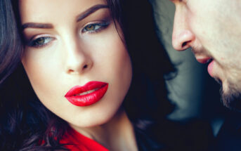 A woman with dark hair and red lipstick looks away thoughtfully while a man leans in close to whisper something, partially visible. The scene conveys an intimate and possibly intense moment between the two.