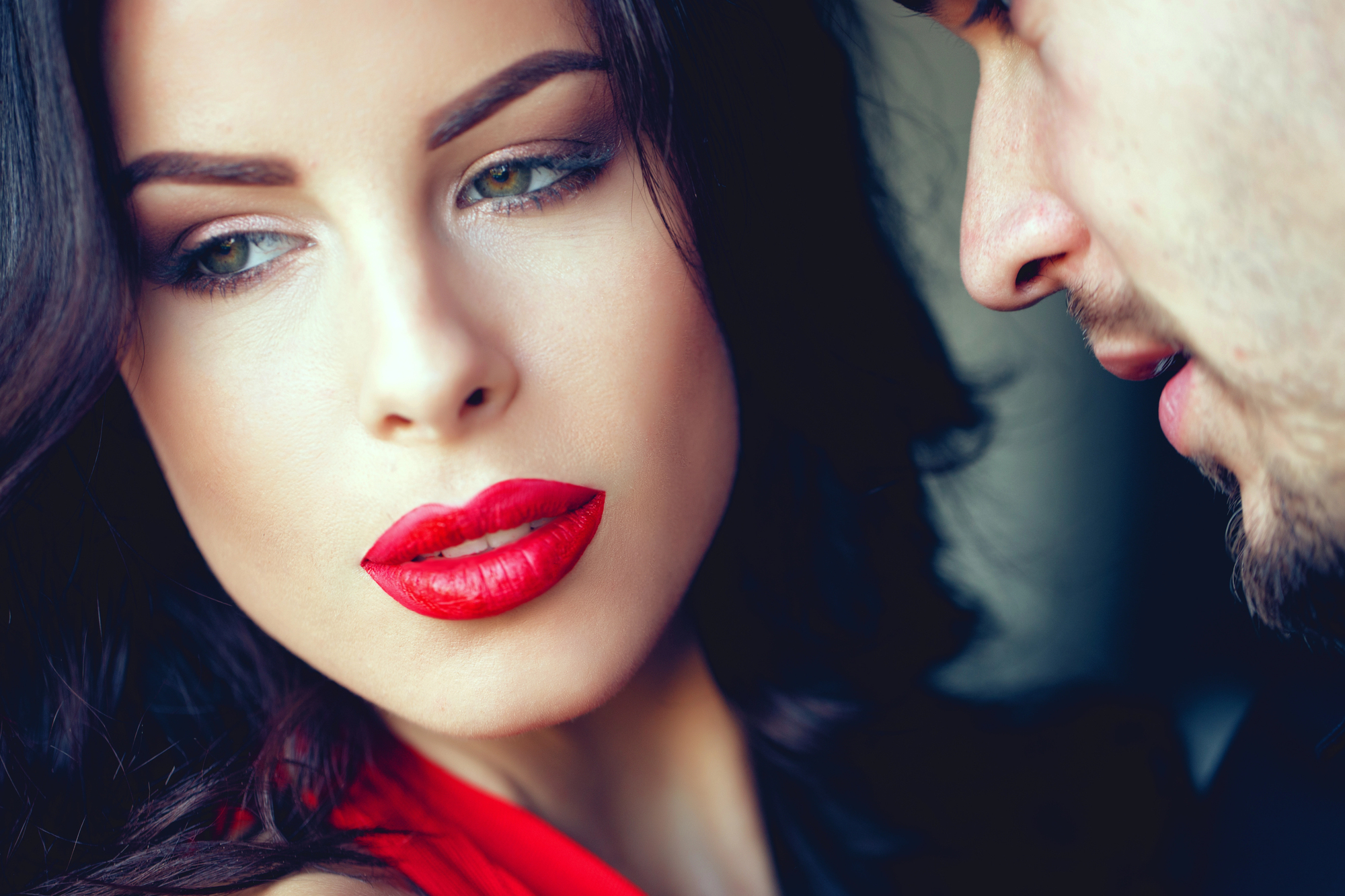 A woman with dark hair and red lipstick looks away thoughtfully while a man leans in close to whisper something, partially visible. The scene conveys an intimate and possibly intense moment between the two.