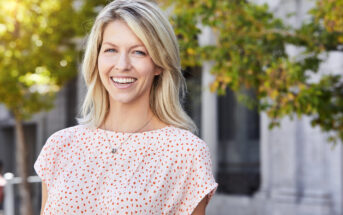 A woman with blonde hair smiles outdoors. She is wearing a light-colored blouse with a red dotted pattern. Green trees and a building are blurred in the background, suggesting a sunny day.