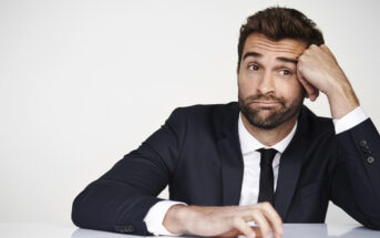 A man with a beard, wearing a dark suit and tie, sits at a table with a neutral expression. His head rests on his right hand, fingers touching his temple. The background is a plain white wall.