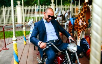 A man in a suit and sunglasses playfully poses on a motorbike ride at a carousel, surrounded by painted animal figures like a giraffe and a horse. The background displays a fence and trees, suggesting an amusement park setting.