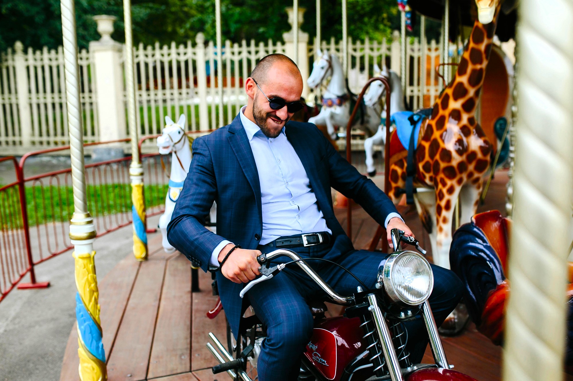 A man in a suit and sunglasses playfully poses on a motorbike ride at a carousel, surrounded by painted animal figures like a giraffe and a horse. The background displays a fence and trees, suggesting an amusement park setting.