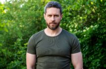 A man with a beard and short hair stands outdoors in front of lush green foliage, wearing a green t-shirt. He looks directly at the camera with a neutral expression.