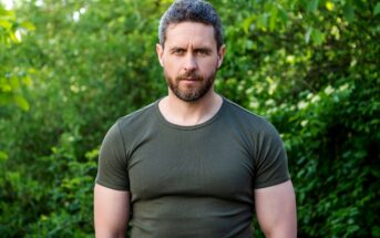 A man with a beard and short hair stands outdoors in front of lush green foliage, wearing a green t-shirt. He looks directly at the camera with a neutral expression.