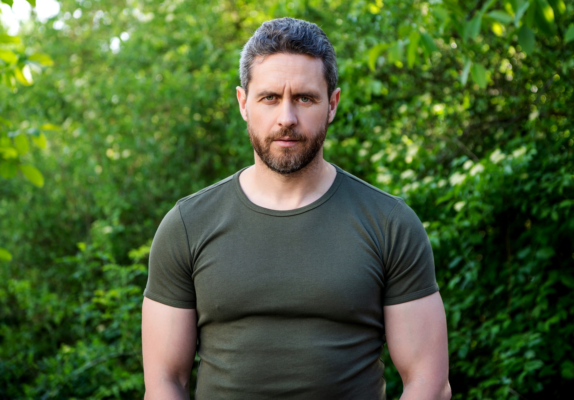 A man with a beard and short hair stands outdoors in front of lush green foliage, wearing a green t-shirt. He looks directly at the camera with a neutral expression.