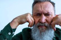 An older man with a long white beard and gray hair appears thoughtful, resting his chin on his fists. He wears a dark green shirt with buttoned cuffs, set against a light blue background. His expression is contemplative.