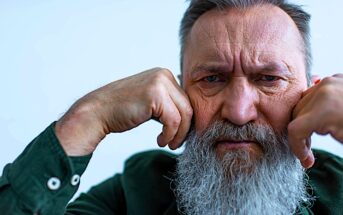 An older man with a long white beard and gray hair appears thoughtful, resting his chin on his fists. He wears a dark green shirt with buttoned cuffs, set against a light blue background. His expression is contemplative.