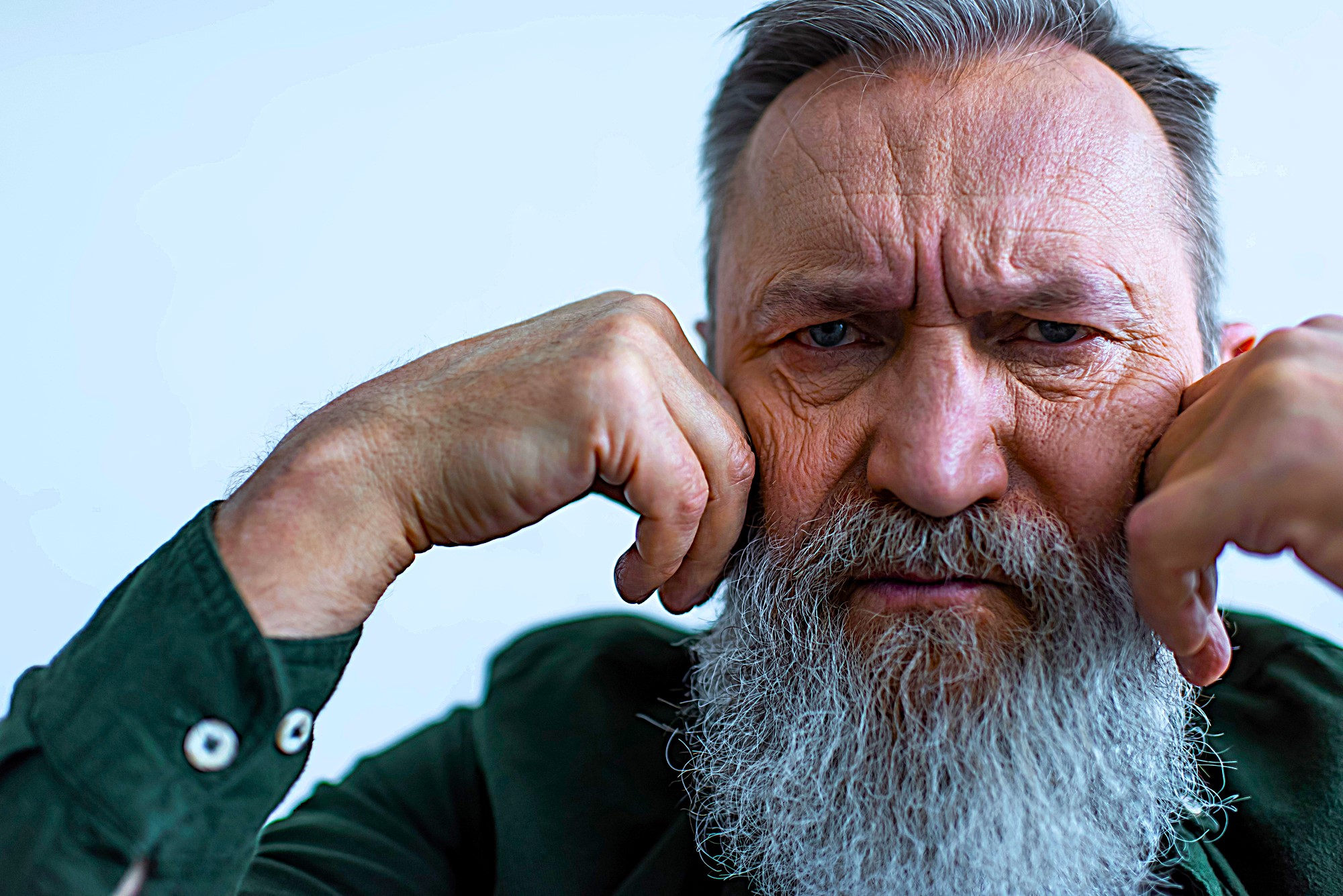 An older man with a long white beard and gray hair appears thoughtful, resting his chin on his fists. He wears a dark green shirt with buttoned cuffs, set against a light blue background. His expression is contemplative.