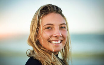 A woman with long blonde hair smiles at the camera. She is outdoors with a blurred background of a beach and ocean, suggesting a warm and sunny day.