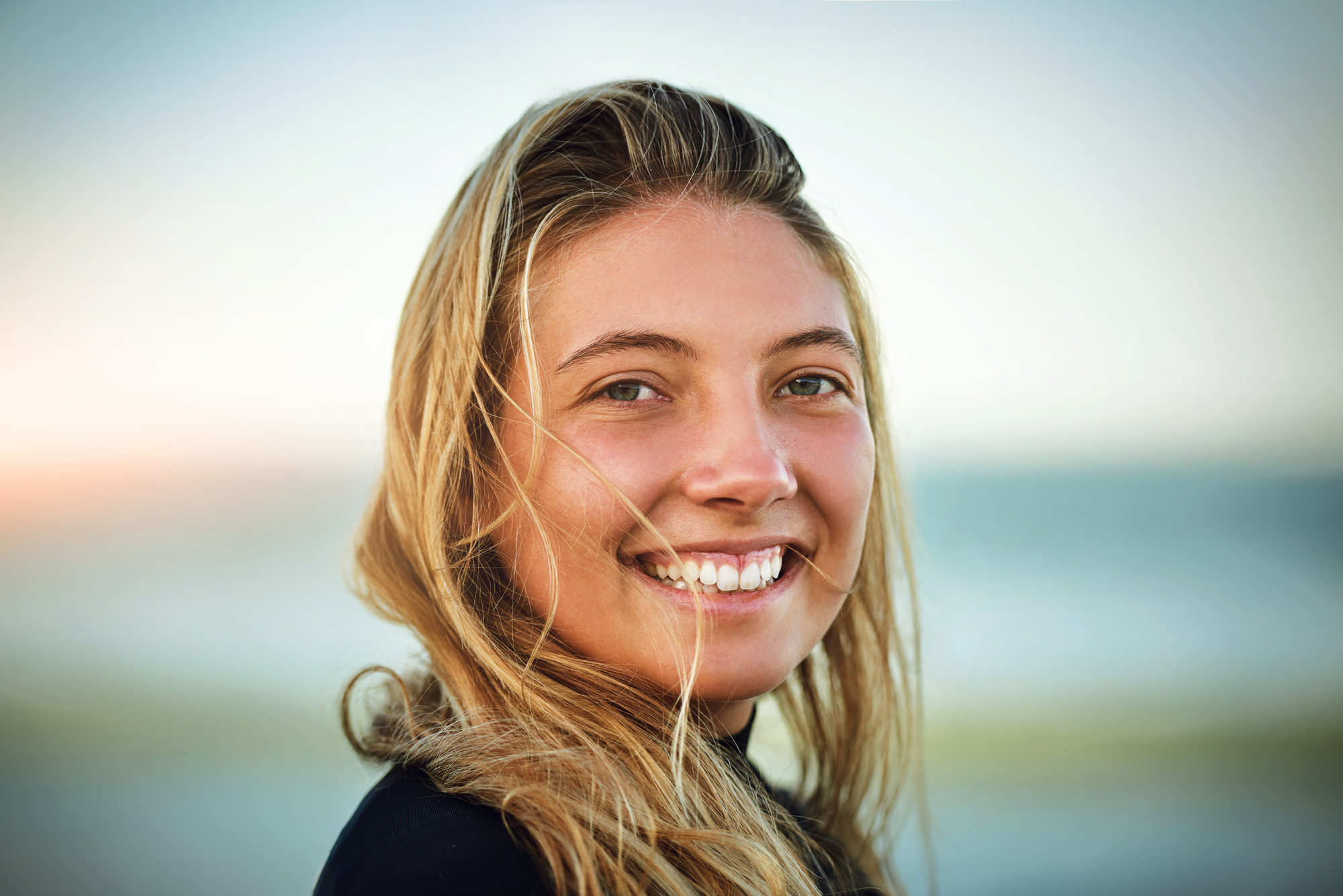 A woman with long blonde hair smiles at the camera. She is outdoors with a blurred background of a beach and ocean, suggesting a warm and sunny day.