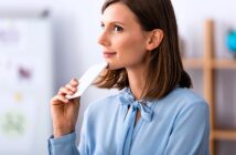 A woman in a light blue blouse holds a white bar up to her chin, looking thoughtful. She stands in a softly lit room with a blurred background, including a shelf and wall decor.