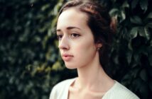 A woman with fair skin and dark hair pulled back is shown in profile against a background of lush green foliage. She has a calm expression, soft features, and is wearing a simple white top.