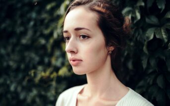 A woman with fair skin and dark hair pulled back is shown in profile against a background of lush green foliage. She has a calm expression, soft features, and is wearing a simple white top.