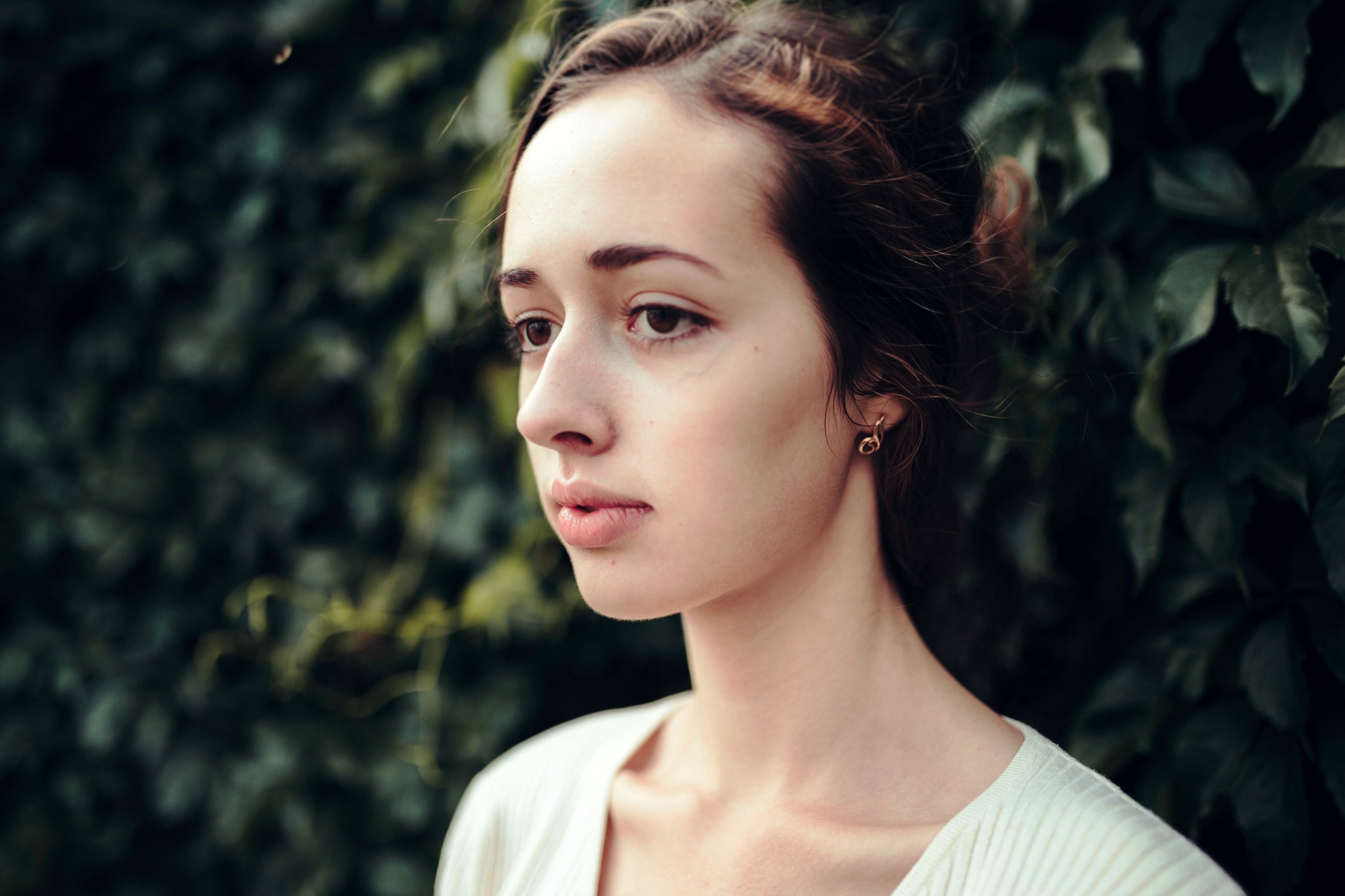 A woman with fair skin and dark hair pulled back is shown in profile against a background of lush green foliage. She has a calm expression, soft features, and is wearing a simple white top.