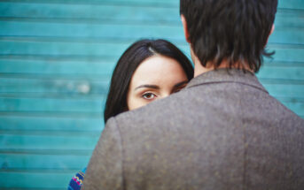 A woman with long dark hair looks past a man's shoulder, with only her eyes and part of her face visible. The man faces away, wearing a brown textured jacket. The background features teal horizontal stripes.