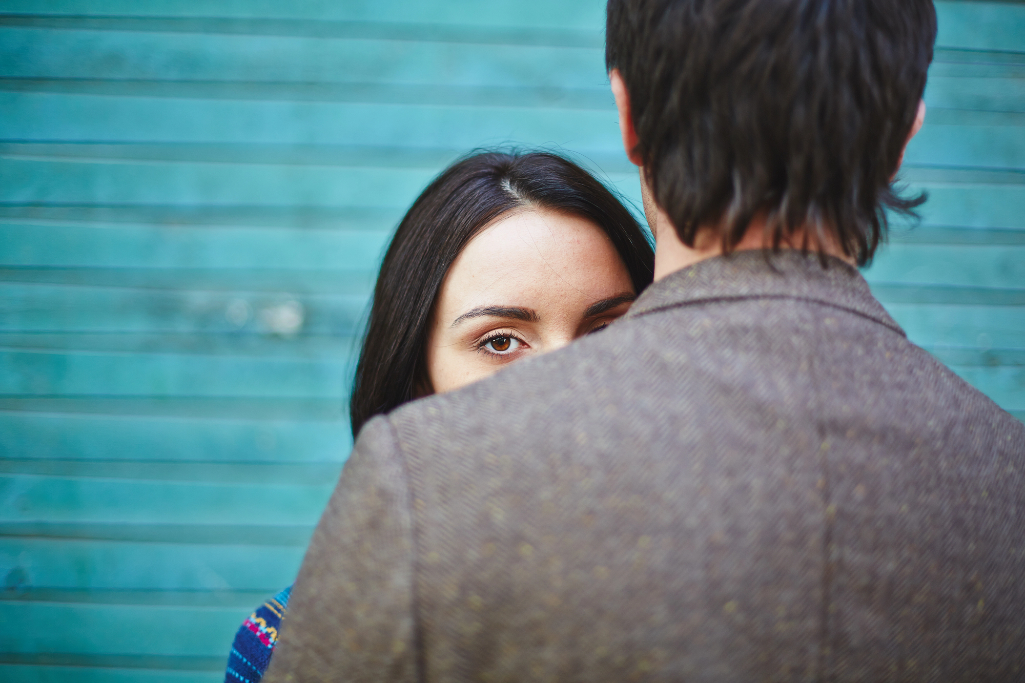 A woman with long dark hair looks past a man's shoulder, with only her eyes and part of her face visible. The man faces away, wearing a brown textured jacket. The background features teal horizontal stripes.