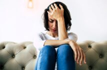 A woman sits on a beige tufted sofa, leaning forward with her hand on her forehead. She has short dark hair and wears a white long-sleeve shirt and blue jeans. Her expression appears stressed or contemplative against a plain white background.