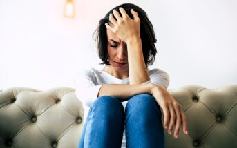 A woman sits on a beige tufted sofa, leaning forward with her hand on her forehead. She has short dark hair and wears a white long-sleeve shirt and blue jeans. Her expression appears stressed or contemplative against a plain white background.