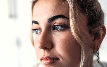 Close-up of a person with light hair and subtle makeup, gazing to the side. The background is softly blurred, highlighting their facial features and expression.