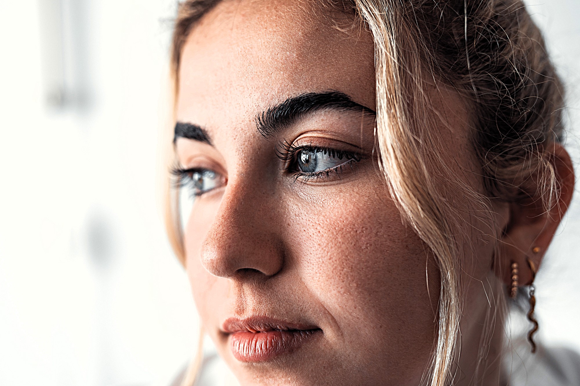 Close-up of a person with light hair and subtle makeup, gazing to the side. The background is softly blurred, highlighting their facial features and expression.