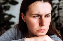 A woman with long brown hair and a grey shirt appears contemplative, resting her chin on her hand. She is gazing off to the side with a serious expression, and there are blurred green plants in the background.