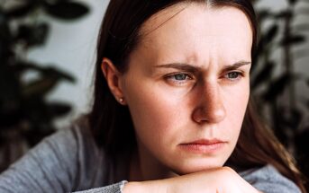 A woman with long brown hair and a grey shirt appears contemplative, resting her chin on her hand. She is gazing off to the side with a serious expression, and there are blurred green plants in the background.