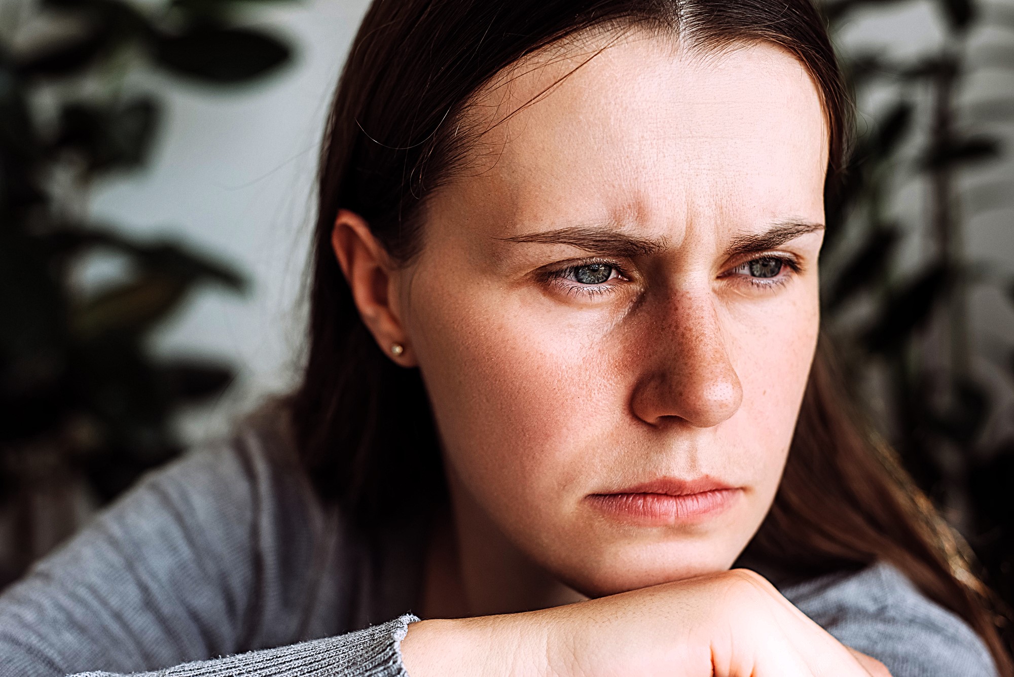 A woman with long brown hair and a grey shirt appears contemplative, resting her chin on her hand. She is gazing off to the side with a serious expression, and there are blurred green plants in the background.