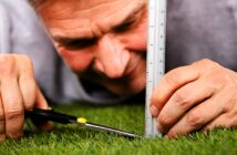 A person lying on grass measures its height with a ruler while precisely trimming it with scissors. The close-up view emphasizes attention to detail and meticulousness.