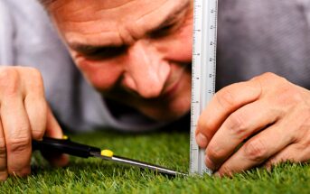 A person lying on grass measures its height with a ruler while precisely trimming it with scissors. The close-up view emphasizes attention to detail and meticulousness.