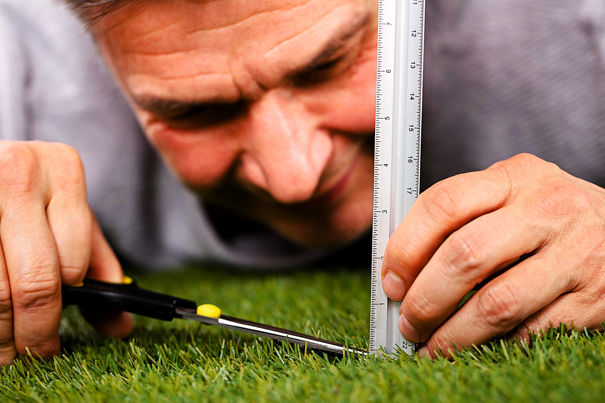 A person lying on grass measures its height with a ruler while precisely trimming it with scissors. The close-up view emphasizes attention to detail and meticulousness.