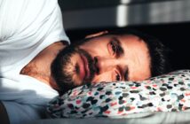 A man with a beard lies on his side on a patterned pillow, wearing a white shirt. He is gazing intently at the camera, partially lit by sunlight, casting shadows around him.