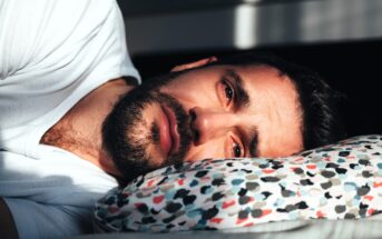 A man with a beard lies on his side on a patterned pillow, wearing a white shirt. He is gazing intently at the camera, partially lit by sunlight, casting shadows around him.