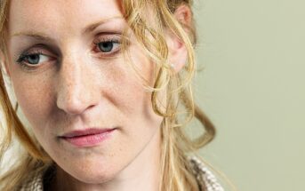 A woman with curly blonde hair and freckles is looking downward with a pensive expression. She is wearing a checkered shirt, and the background is a soft green color.