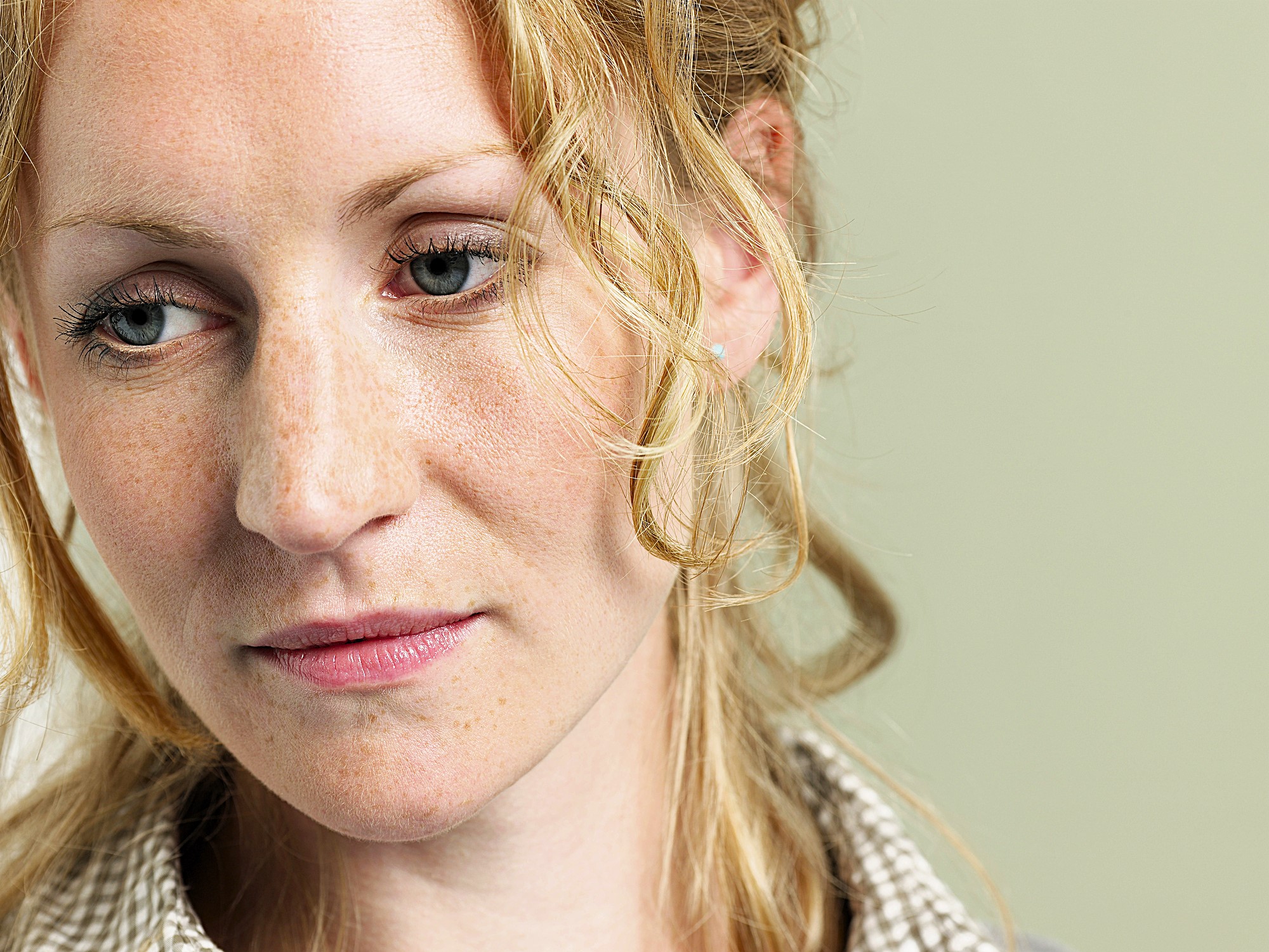 A woman with curly blonde hair and freckles is looking downward with a pensive expression. She is wearing a checkered shirt, and the background is a soft green color.