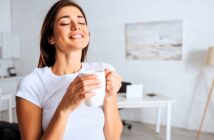 A woman in a white shirt stands in a bright room, eyes closed, smiling while holding a white mug. A desk and a painting on the wall are visible in the blurred background.