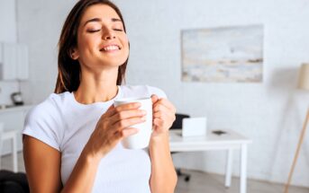 A woman in a white shirt stands in a bright room, eyes closed, smiling while holding a white mug. A desk and a painting on the wall are visible in the blurred background.