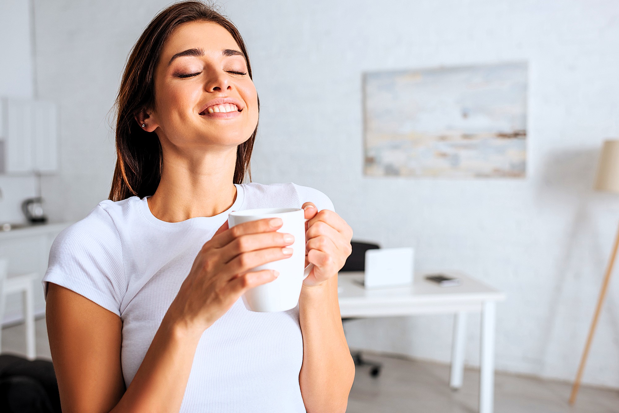 A woman in a white shirt stands in a bright room, eyes closed, smiling while holding a white mug. A desk and a painting on the wall are visible in the blurred background.