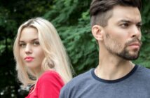 A woman with long blonde hair and a red top stands next to a man with short dark hair, a beard, and a gray t-shirt. The woman faces forward while the man looks off into the distance. Both are outdoors with lush green foliage in the background.