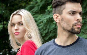 A woman with long blonde hair and a red top stands next to a man with short dark hair, a beard, and a gray t-shirt. The woman faces forward while the man looks off into the distance. Both are outdoors with lush green foliage in the background.