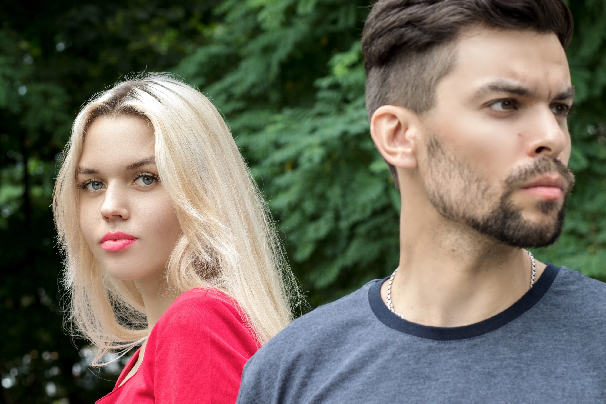 A woman with long blonde hair and a red top stands next to a man with short dark hair, a beard, and a gray t-shirt. The woman faces forward while the man looks off into the distance. Both are outdoors with lush green foliage in the background.