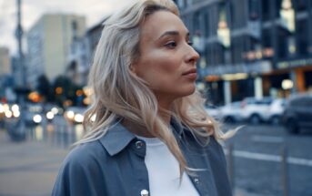 A person with long, wavy blonde hair looks to the side while standing on a city street. They wear a white top with a blue-gray jacket. The background features blurred buildings and cars on a busy road.