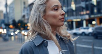 A person with long, wavy blonde hair looks to the side while standing on a city street. They wear a white top with a blue-gray jacket. The background features blurred buildings and cars on a busy road.