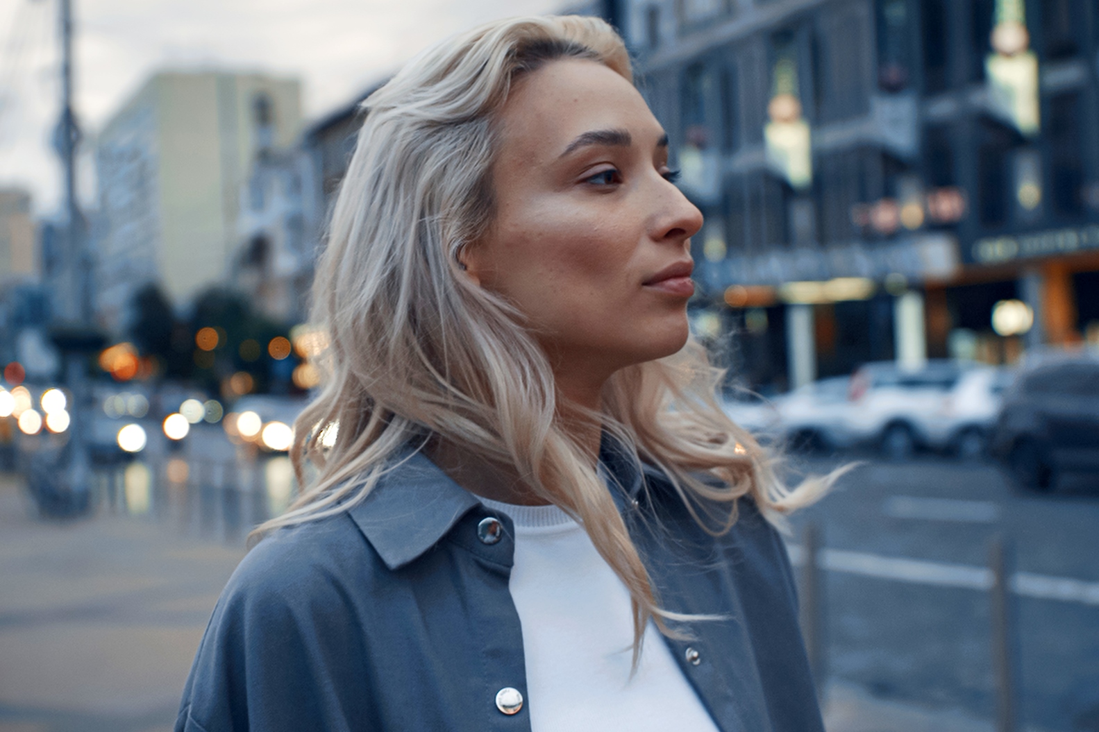 A person with long, wavy blonde hair looks to the side while standing on a city street. They wear a white top with a blue-gray jacket. The background features blurred buildings and cars on a busy road.