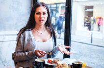 A woman with long dark hair, wearing a light gray cardigan and white top, sits at a table in a café. She looks surprised and holds a fork. The table has several dishes, including fruit, coffee, and croissants. A window showing a street view is behind her.