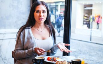 A woman with long dark hair, wearing a light gray cardigan and white top, sits at a table in a café. She looks surprised and holds a fork. The table has several dishes, including fruit, coffee, and croissants. A window showing a street view is behind her.